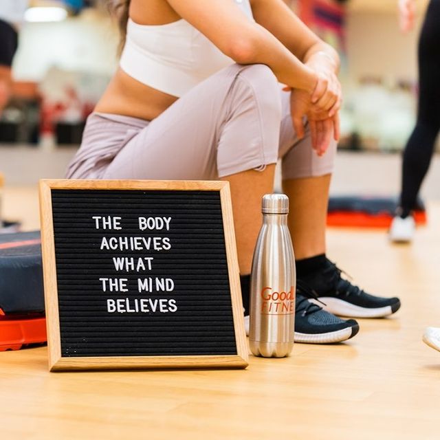 Man in grey shirt does standing shoulder presses with dumbbells while a second man on a bench in a white t-shirt observes his form