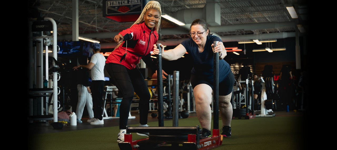A GoodLife Fitness Personal Trainer directing a member who is pushing a fitness sled on turf.