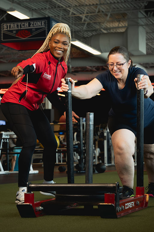 A GoodLife Fitness Personal Trainer directing a member who is pushing a fitness sled on turf.